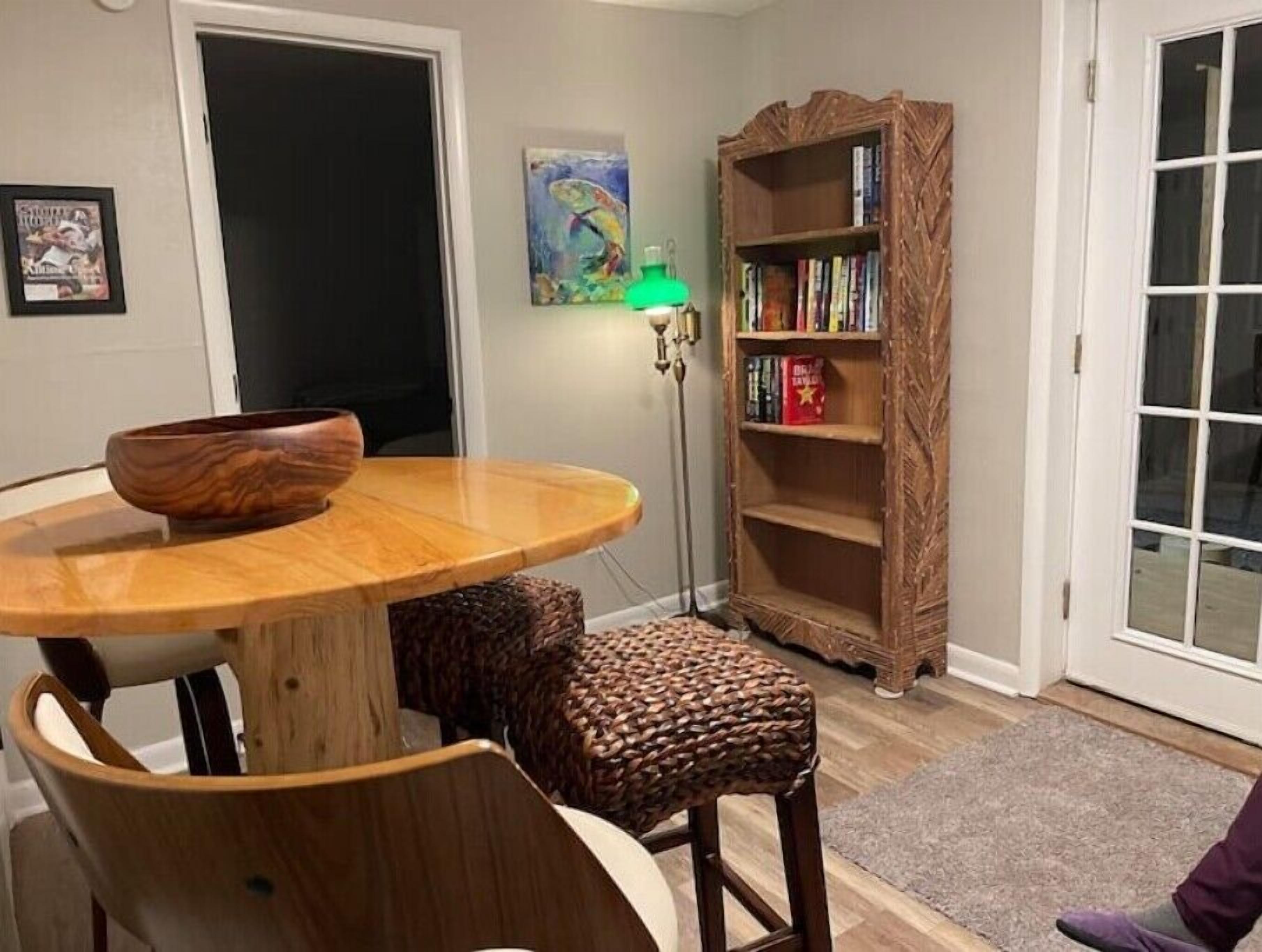 Een eetruimte met een houten tafel, stoelen, een geweven kruk, een boekenkast en een vloerlamp.