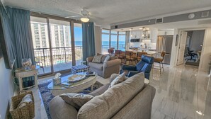 A spacious living room with a sofa, coffee table, and chairs, a kitchen area with modern appliances, and large windows offering a view of high-rise buildings and a balcony.
