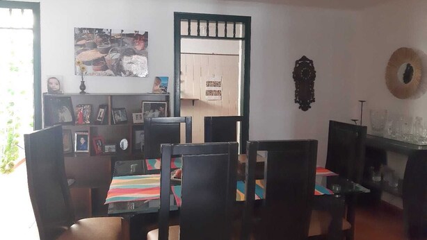Un comedor con una mesa dispuesta para cuatro personas, rodeada de sillas. Hay una estantería con varios objetos y un cuadro enmarcado encima. Un espejo decorativo cuelga en la pared, y se aprecia una puerta con ventana al fondo.