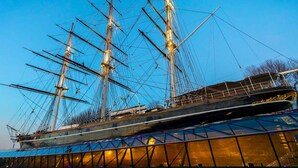 A large ship with multiple masts, docked at a modern building with a glass facade.