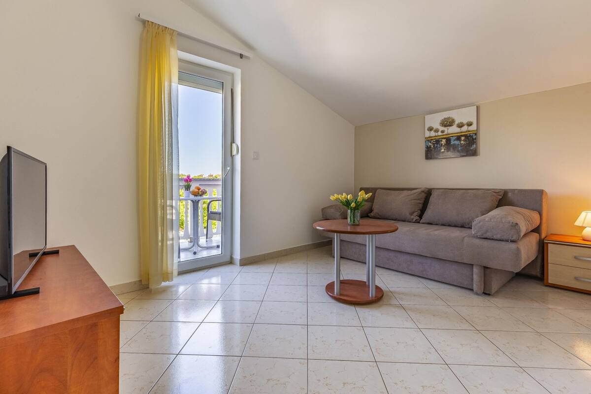 A modern living room with a sofa, a small table with flowers, a TV, and a balcony with yellow curtains.