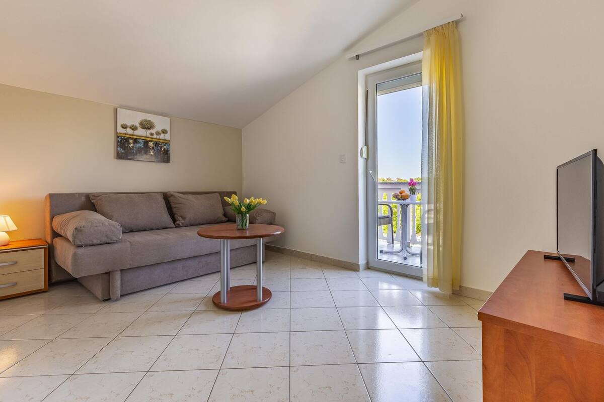 A modern living room with a sofa, a small round table with a vase of flowers, a lamp, a TV, and a balcony with yellow curtains.