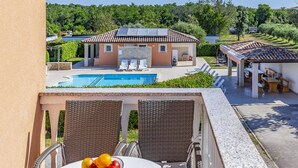 A balcony with wicker chairs and a table with a fruit bowl. In the background, there is a swimming pool, a building with a tiled roof, and a covered area with benches. - Apartments with Shared Pool Rocco 1 (Medulin)