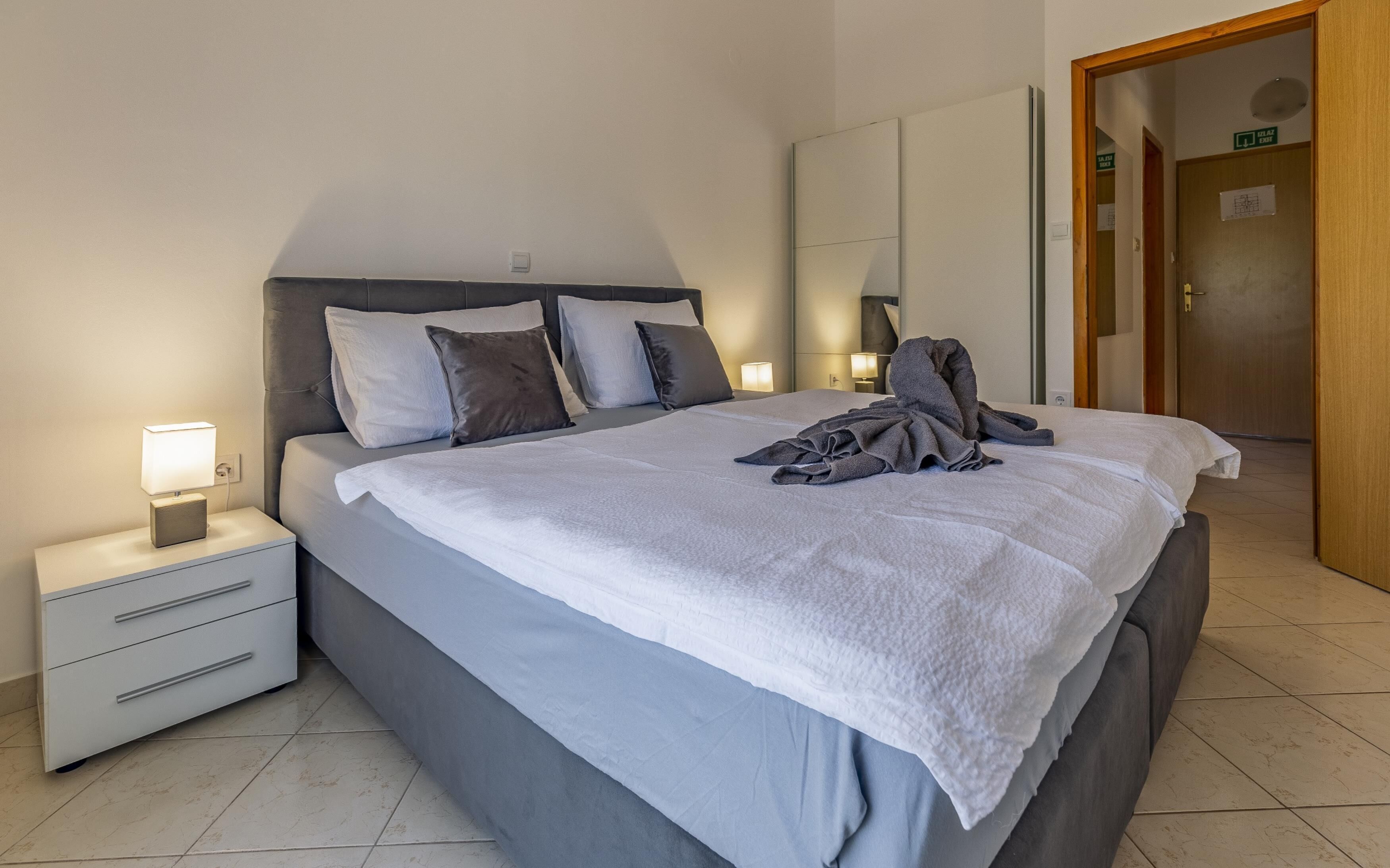 A neatly made bed with a white duvet, grey pillows, and a folded towel on the bed. There is a bedside table with a lamp and a drawer. The room has a wooden door and a white wall with a light switch.