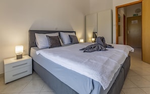 A neatly made bed with a white duvet, grey pillows, and a folded towel on the bed. There is a bedside table with a lamp and a drawer. The room has a wooden door and a white wall with a light switch. - Apartments with Shared Pool Rocco 1 (Medulin)
