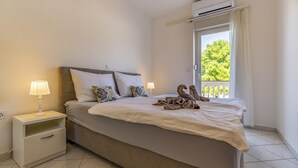 A neatly made bed with a blanket and a decorative item, a bedside table with a lamp, and a window with a view of greenery. - Apartments with Shared Pool Rocco 1 (Medulin)