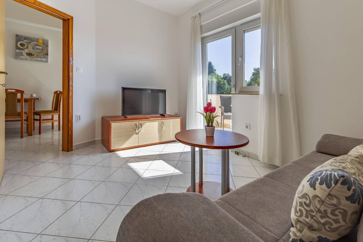 A modern living room with a sofa, a TV on a wooden cabinet, a small round table with a vase of flowers, and a view of the outdoors through large windows.