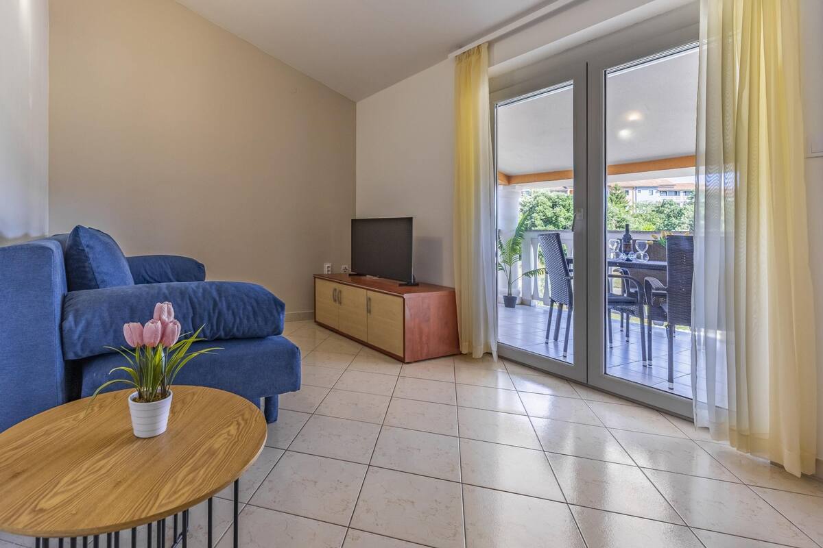 A living room with a sofa, a TV on a wooden cabinet, a small table with a plant, and a sliding glass door leading to an outdoor area with chairs and a table.