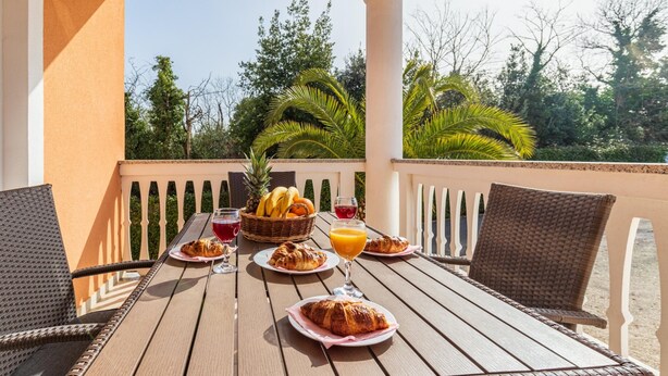 A wooden table set with croissants, fruit, and drinks on an outdoor patio with wicker chairs and a view of trees. - Apartments with Shared Pool Rocco 1 (Medulin)
