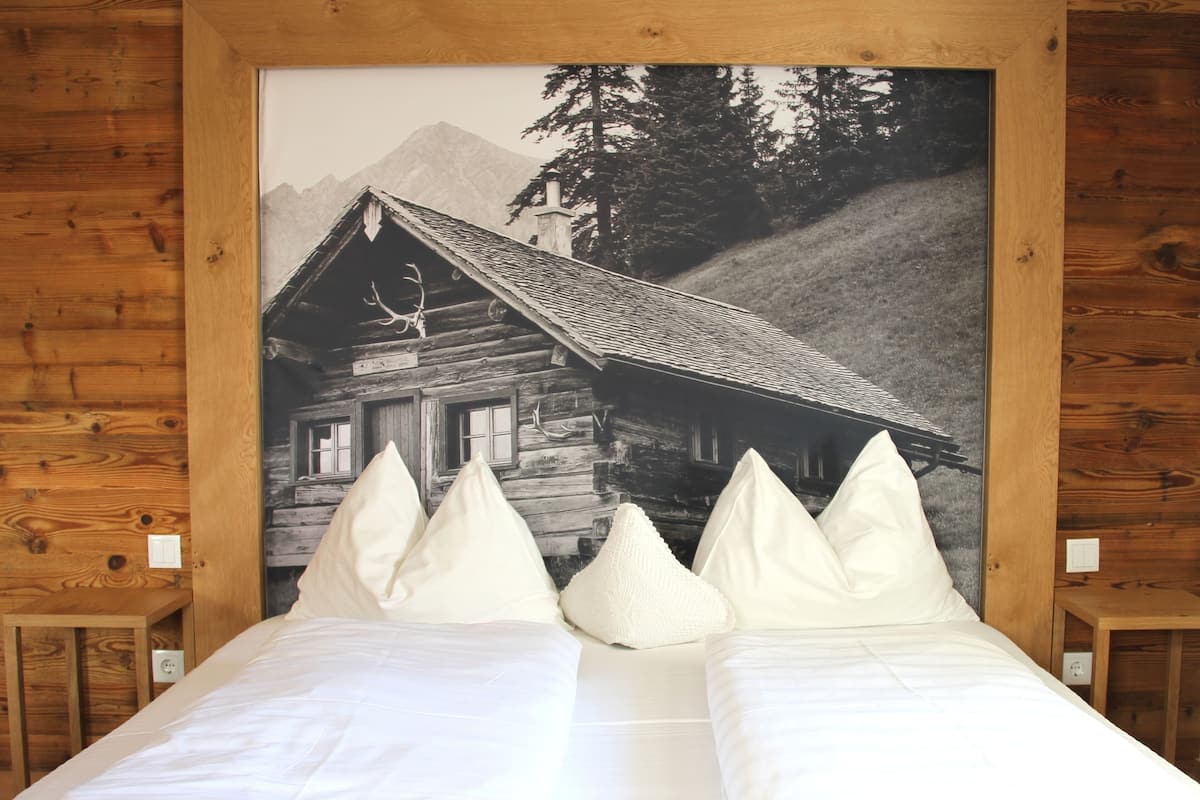 Una cama con ropa de cama y almohadas blancas, enmarcada por un cabecero de madera con una imagen de una cabaña de troncos y montañas al fondo.