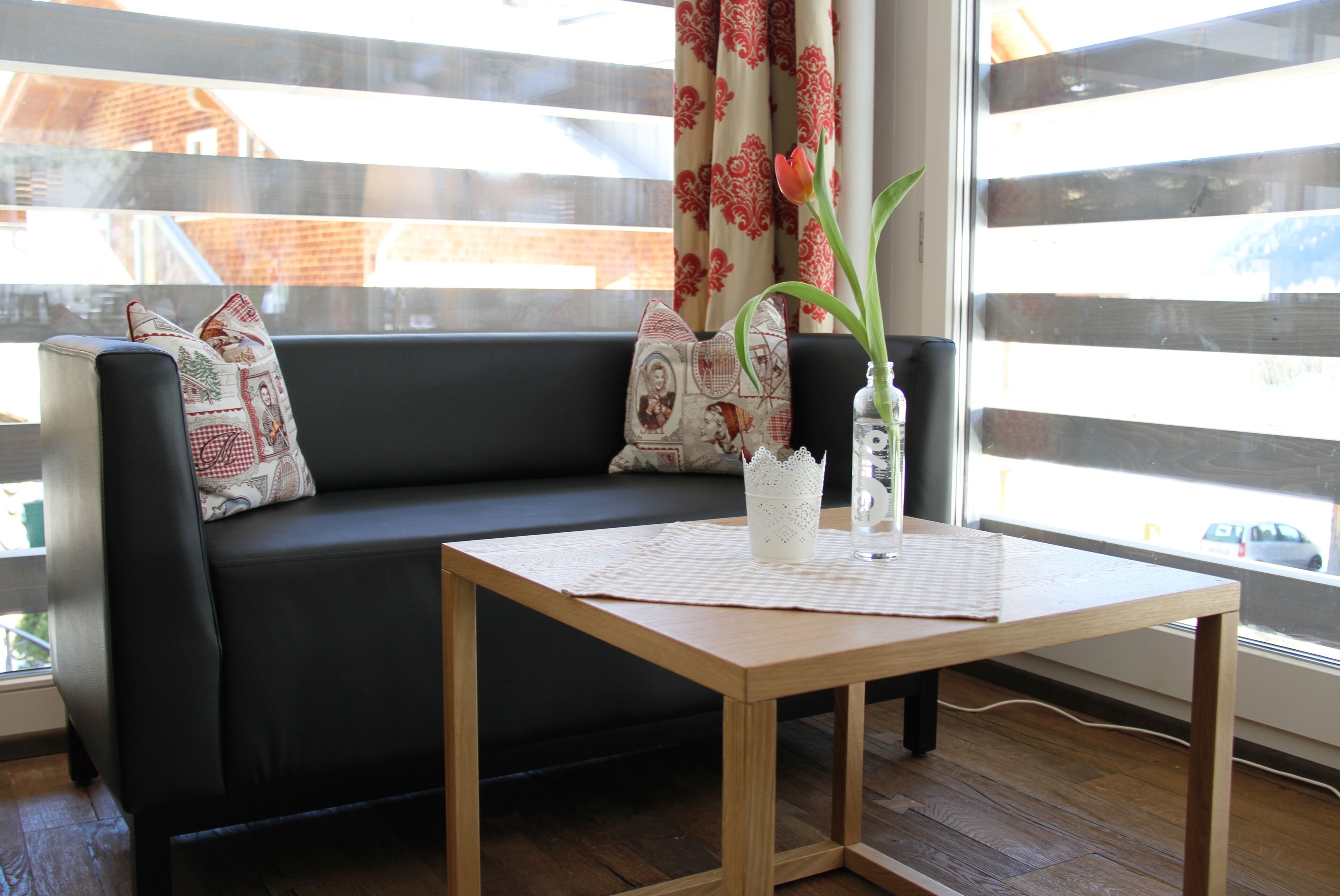 A modern living room with a black leather sofa, patterned cushions, a wooden coffee table, and a vase with a single tulip.
