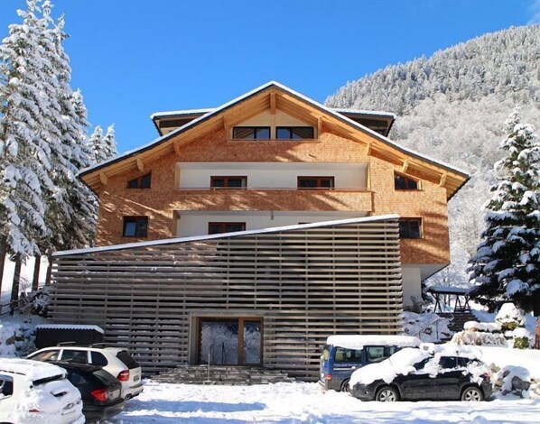A multi-story wooden chalet with a sloped roof, a covered entrance, and a parking area with cars. - Alpen Suites Brandnertal (Brand)