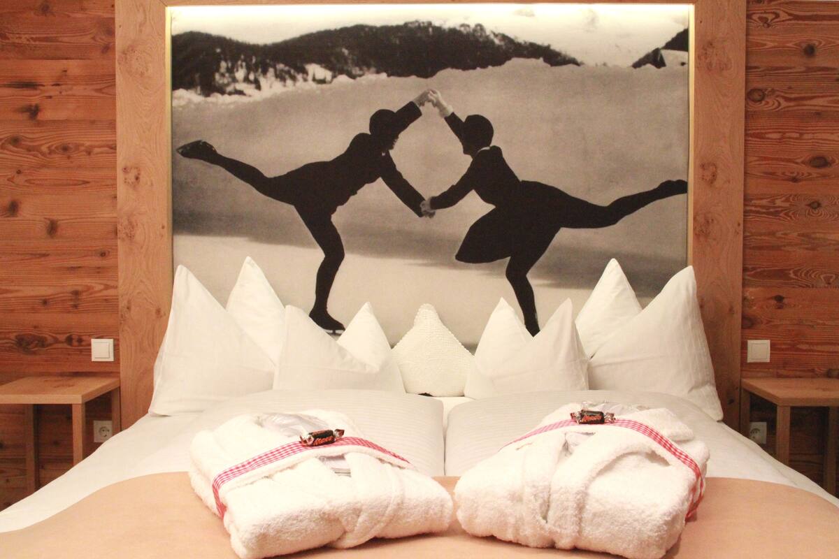 Una cama con un cuadro grande enmarcado de dos personas bailando en un paisaje nevado, almohadas blancas y dos toallas dobladas.