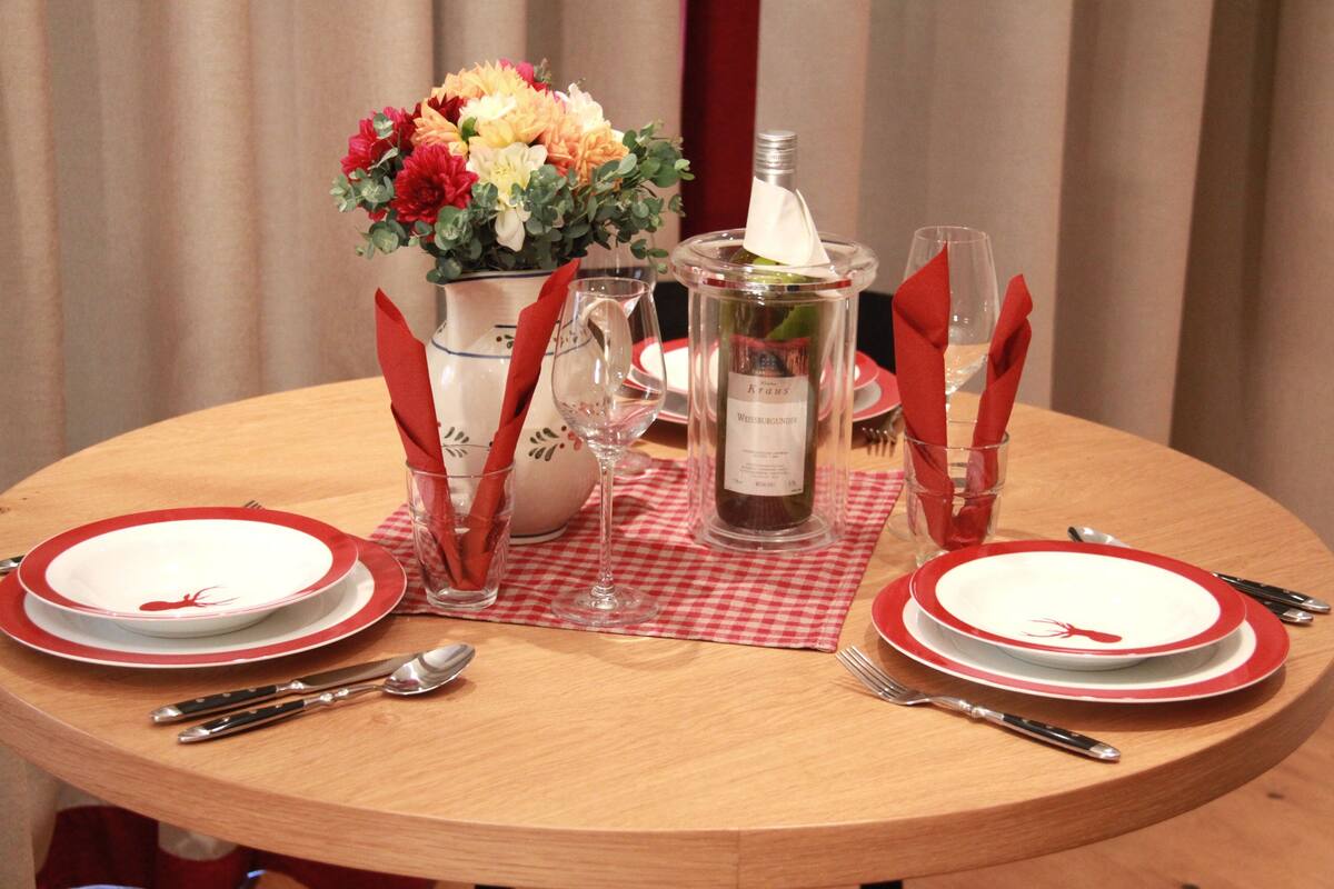 Una mesa redonda para comer con platos blancos y rojos, cubiertos y una botella de vino.