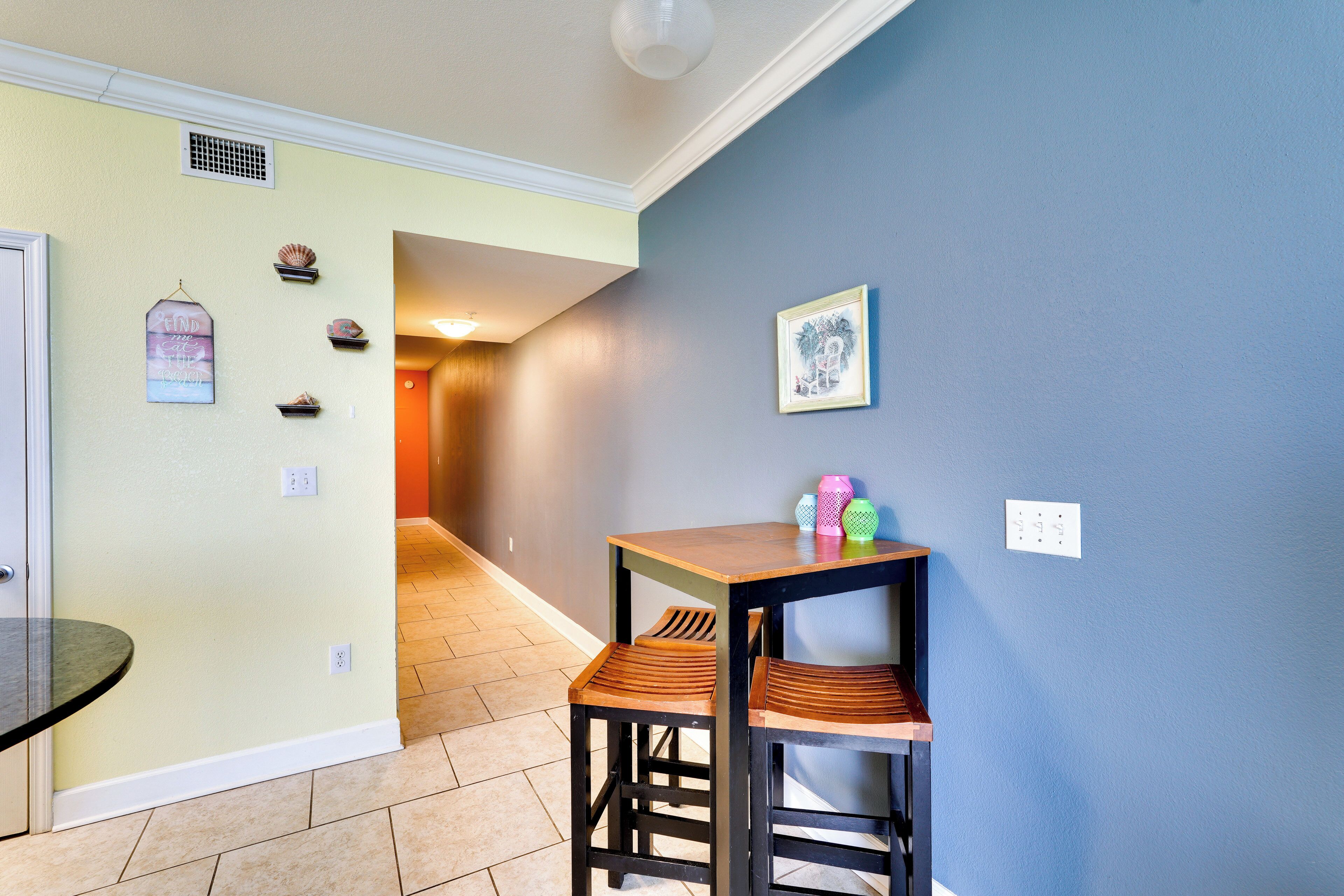 A hallway with a table and two chairs, a vent, and a framed picture on the wall.