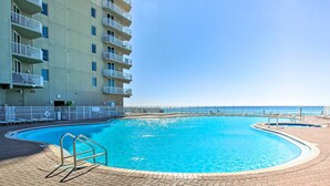 En swimmingpool med kristallklart blått vatten, omgiven av en terrass av tegel, och belägen intill en hög byggnad med balkonger.