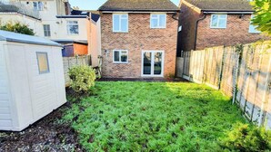 A residential backyard with a grassy lawn, a wooden fence, and a brick house with a front door and windows. - Impeccable 4-bed House in Cheltenham (Cheltenham)