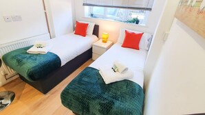 A small, clean room with two single beds, a bedside table with a lamp, and a window with blinds. - Impeccable 4-bed House in Cheltenham (Cheltenham)