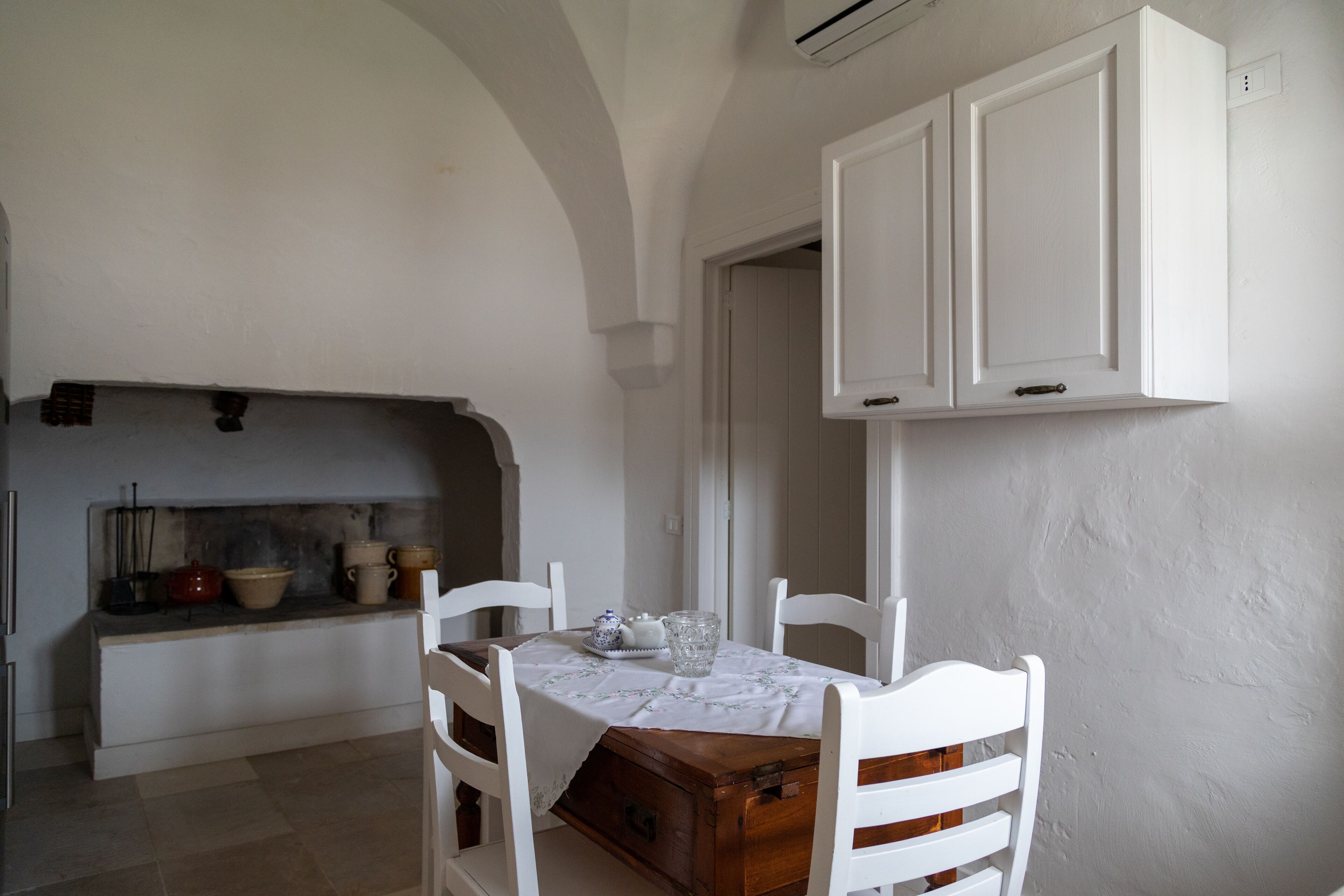 A dining area with a wooden table and white chairs, a white cabinet above, and a kitchen area with pots and pans.