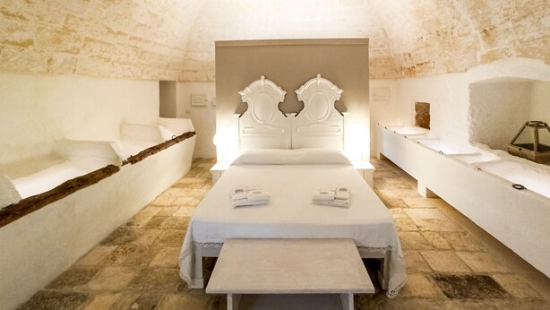 A room with a stone wall, a bed with a white headboard, a bench, and a bedside table with a lamp. - Masseria Scaledda (Manduria)