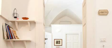 A hallway with white walls, a tiled floor, and a door labeled "LA MACCHIA". Shelves with books and bowls are on the left wall.
