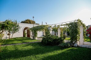 A courtyard with a white building featuring an arched entrance, surrounded by lush green grass and flowering plants. - Masseria Scaledda (Manduria)