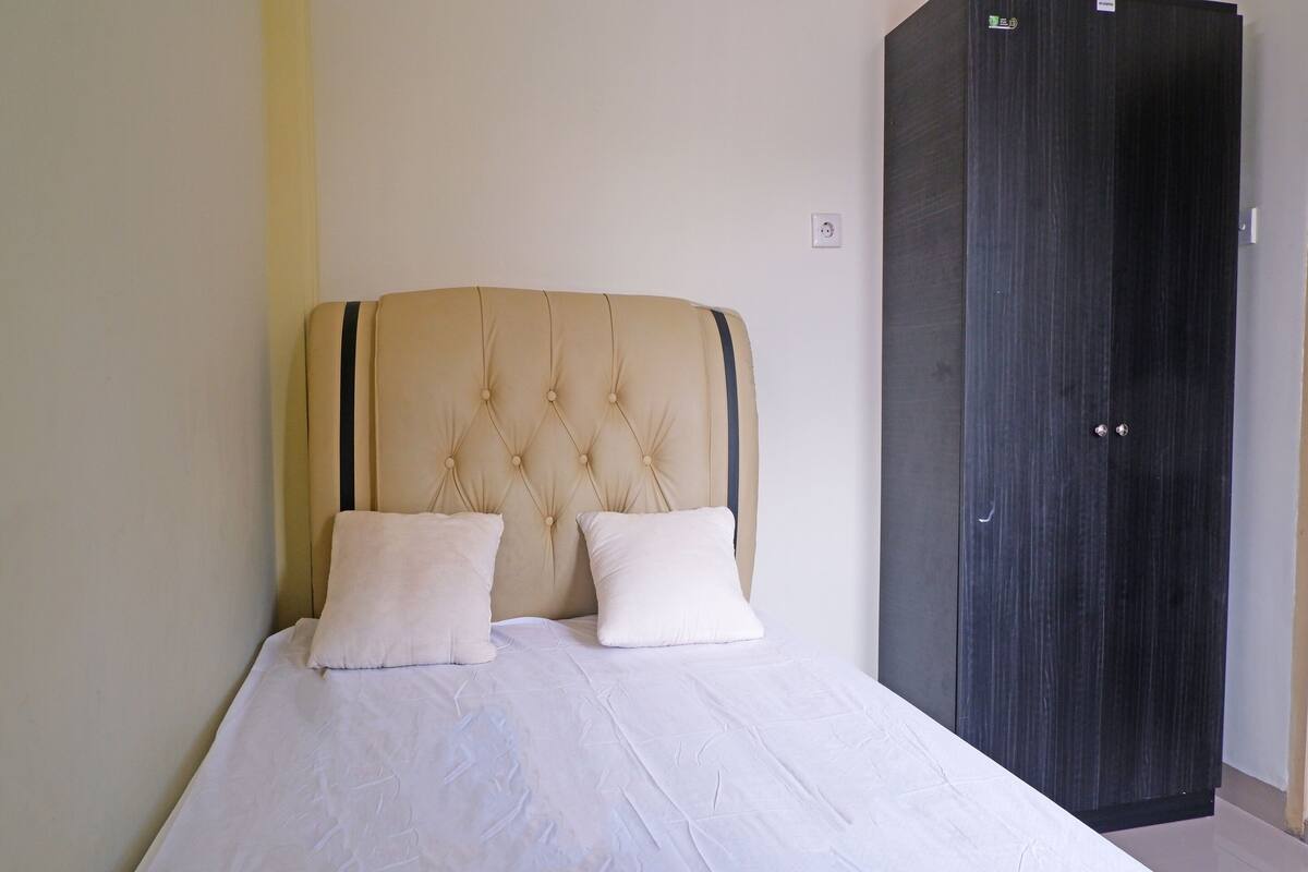 Een bed met een gestoffeerd hoofdbord en twee kussens, een donkere houten garderobe, en een effen muur met een lichtschakelaar.