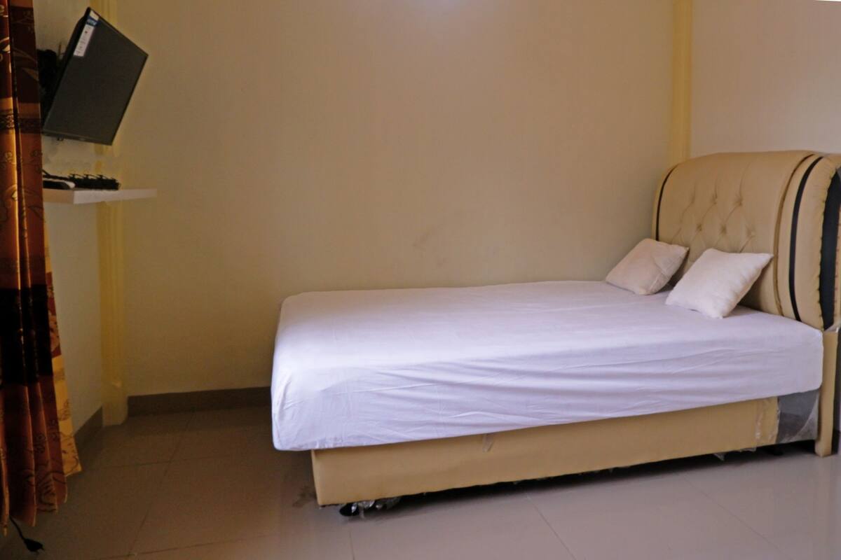 Een enkel bed met hoofdbord, een flatscreen-tv bevestigd aan een plank erboven en een gordijn aan de linkerkant.