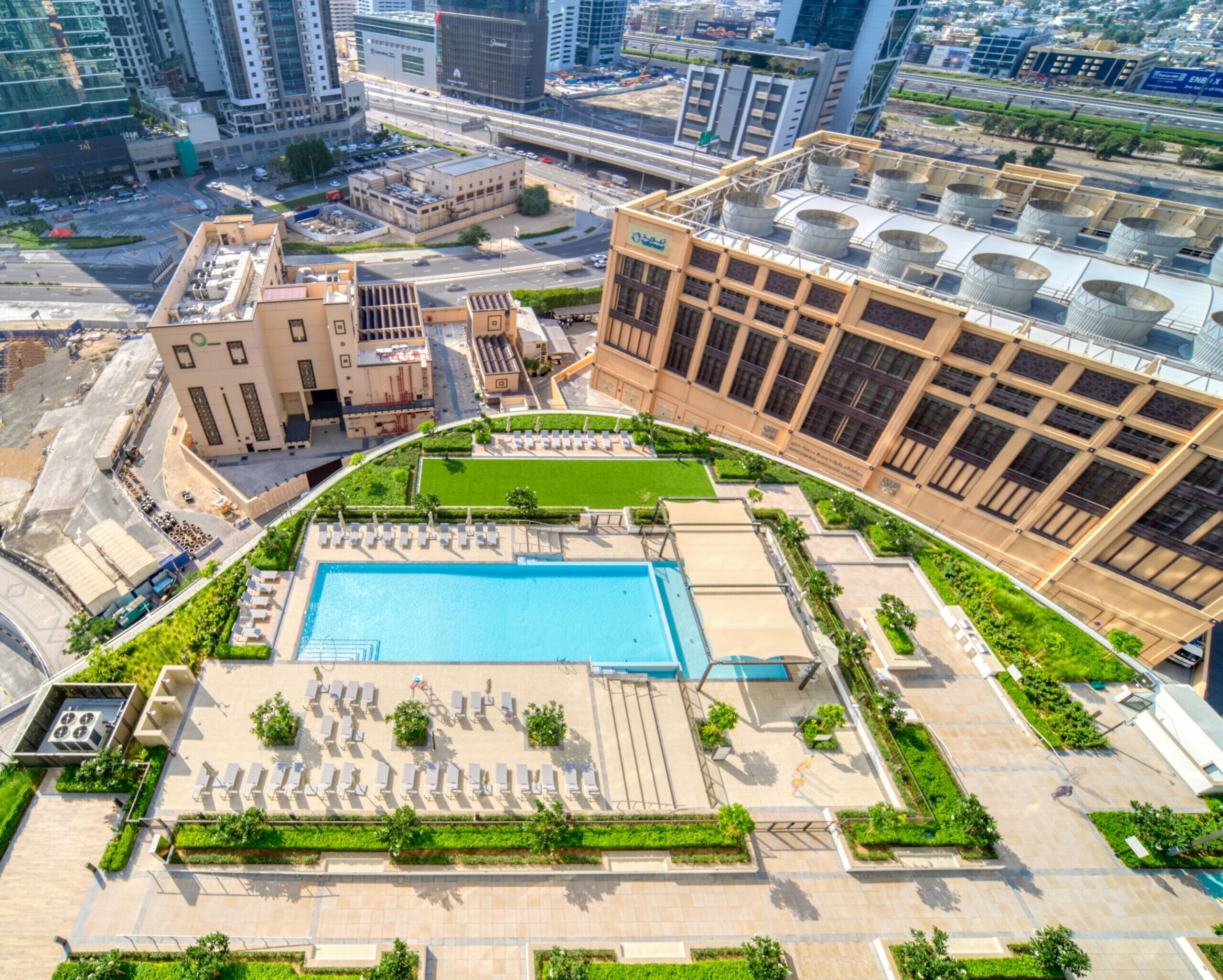 A rooftop swimming pool with sun loungers, surrounded by greenery and adjacent to a modern building with large windows.