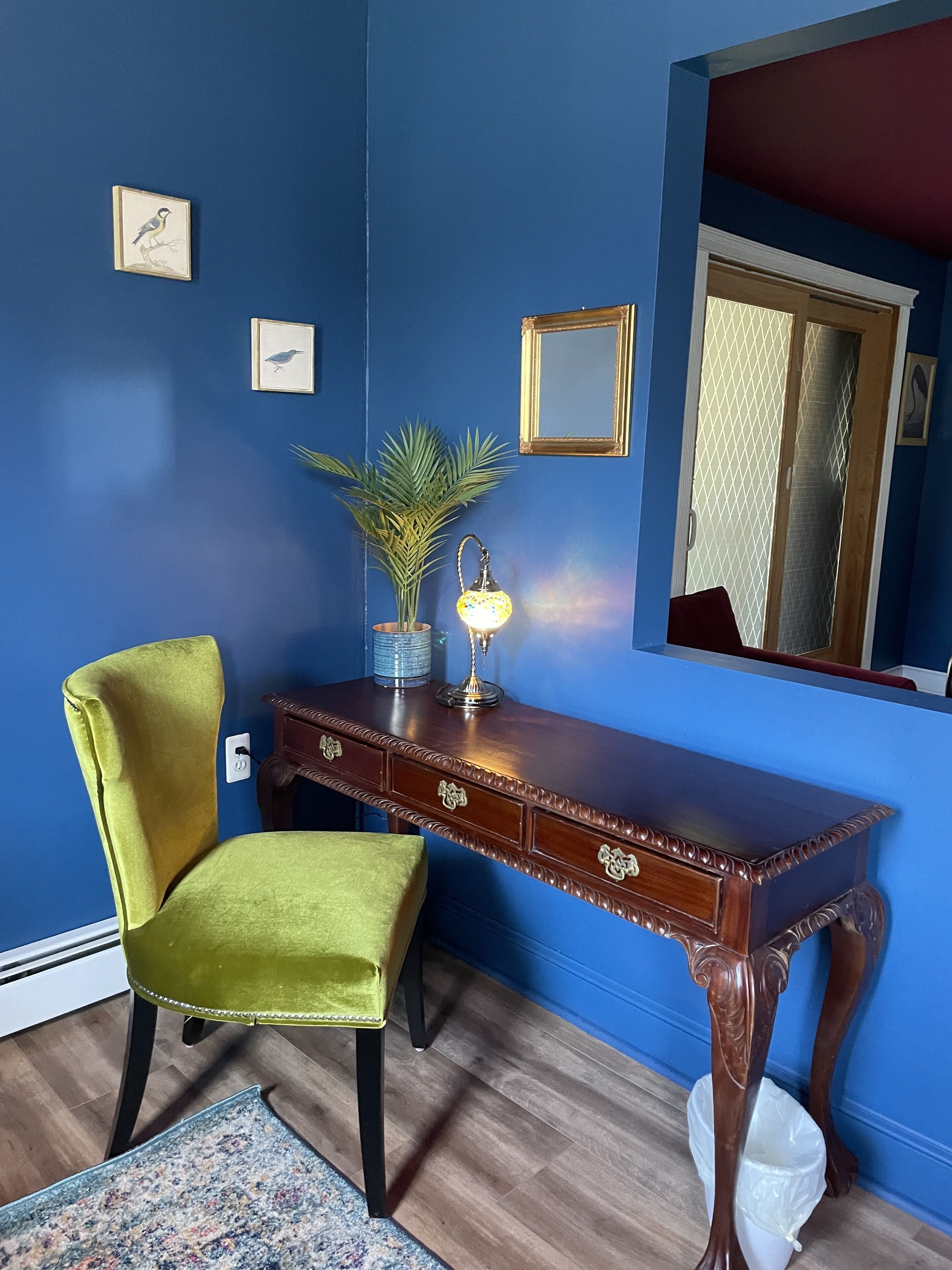 A room with a blue wall, a wooden desk with drawers, a green chair, a small potted plant, a wall-mounted mirror, and a framed picture.