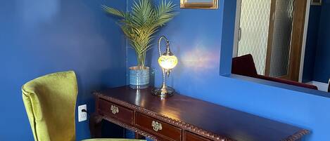 A room with a blue wall, a wooden desk with drawers, a green chair, a small potted plant, a wall-mounted mirror, and a framed picture.