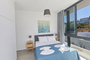 A modern bedroom with a large bed, a painting on the wall, and a view of the outdoors through a large window. - Protaras Elite Pearl Villa Ep4b (Paralimni)