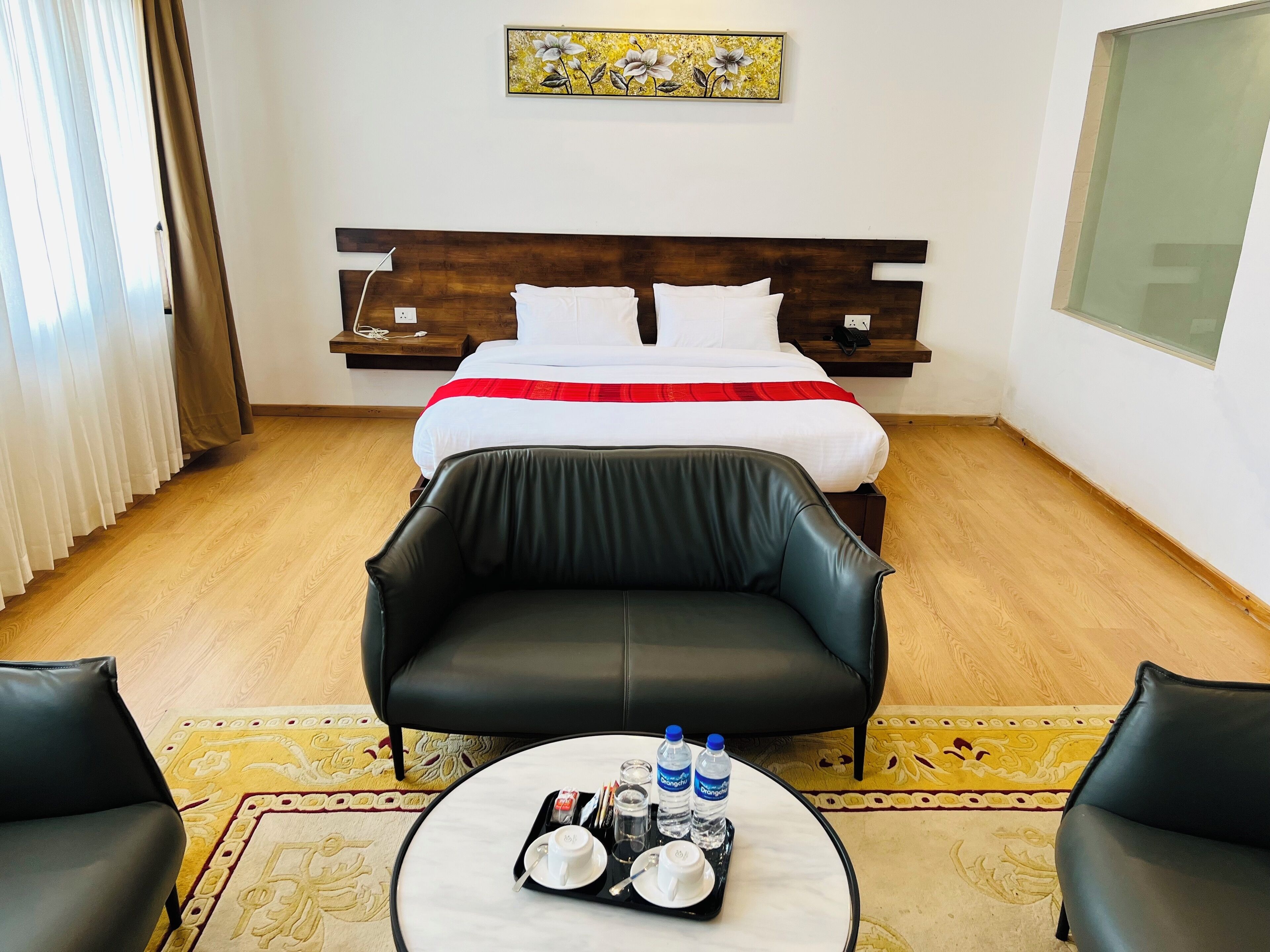 Una habitación de hotel con una cama, un sofá de cuero negro, una mesita redonda con un juego de botellas de agua y vasos, y un cuadro en la pared.