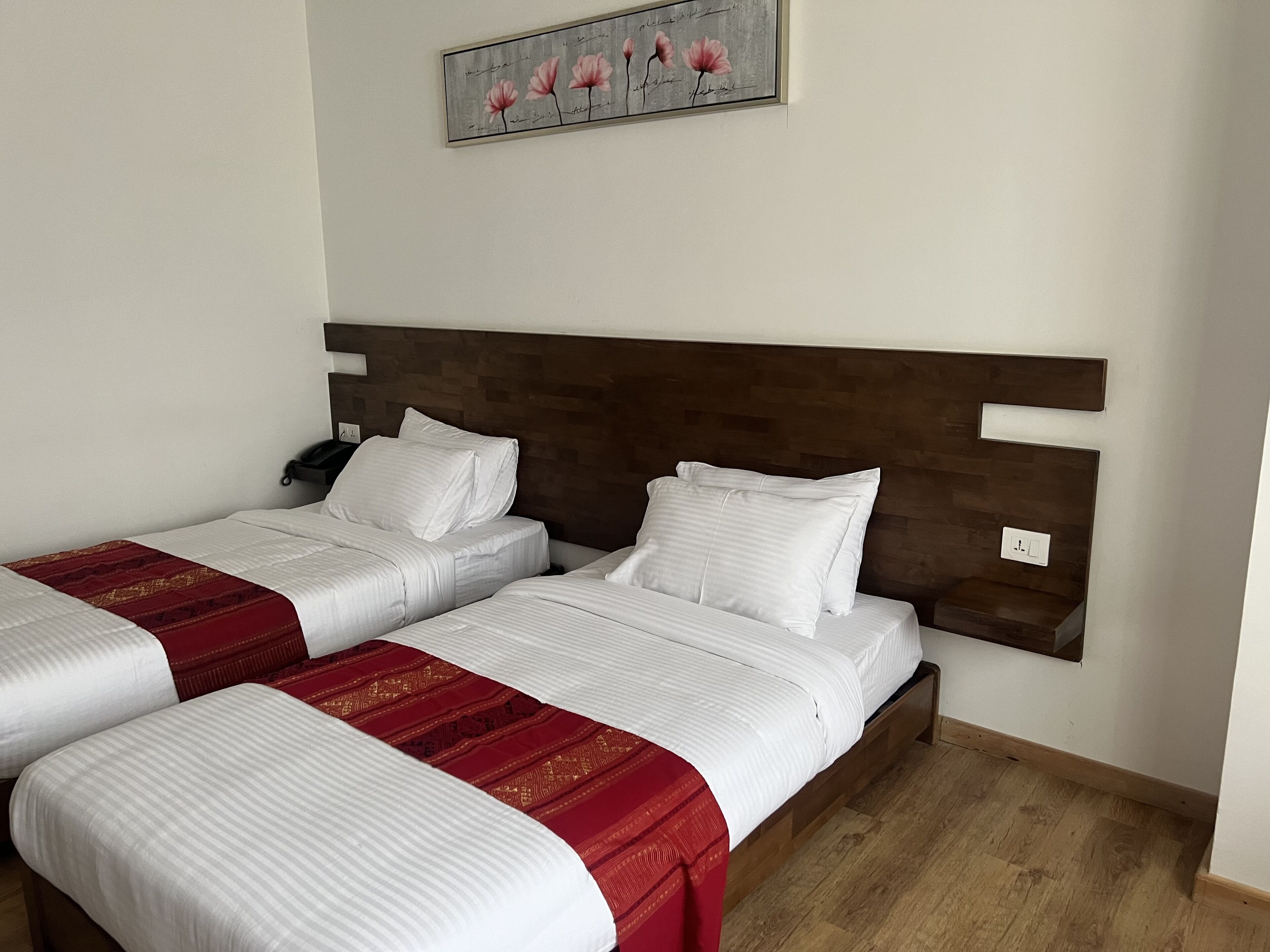 Habitación de hotel con dos camas, cada una con ropa de cama blanca y detalles en rojo, cabeceros de madera y una obra de arte en la pared con flores rosadas.