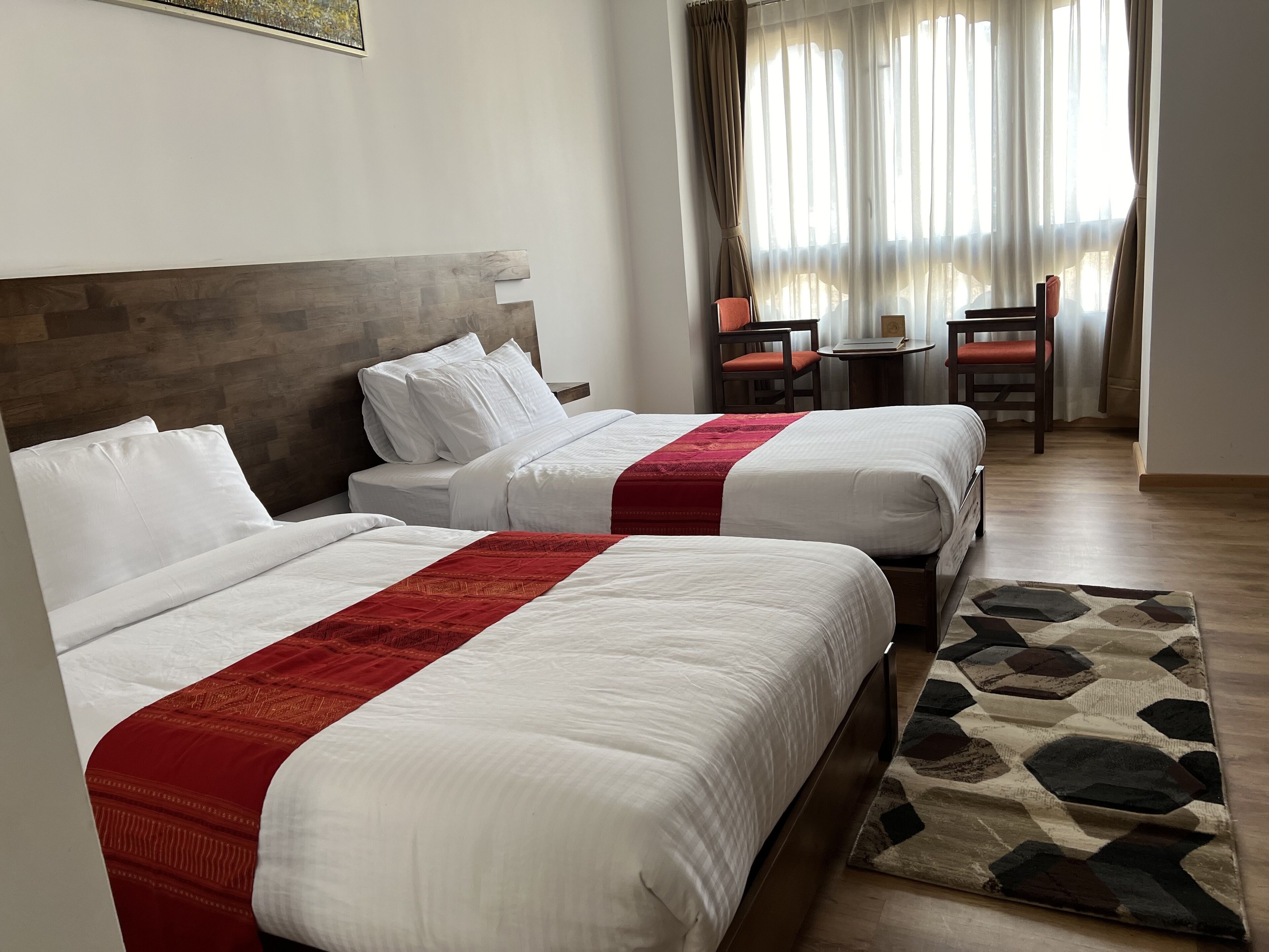 Une chambre d’hôtel avec deux lits, une tête de lit en bois, une petite table avec des chaises et une vue sur l’extérieur à travers des rideaux légers.