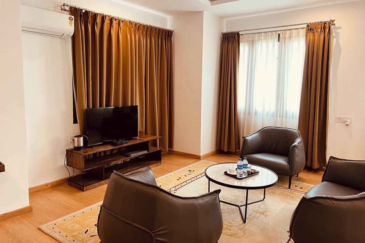 A modern living room with a flat-screen TV on a wooden stand, two leather chairs, a coffee table, and a large window with curtains.