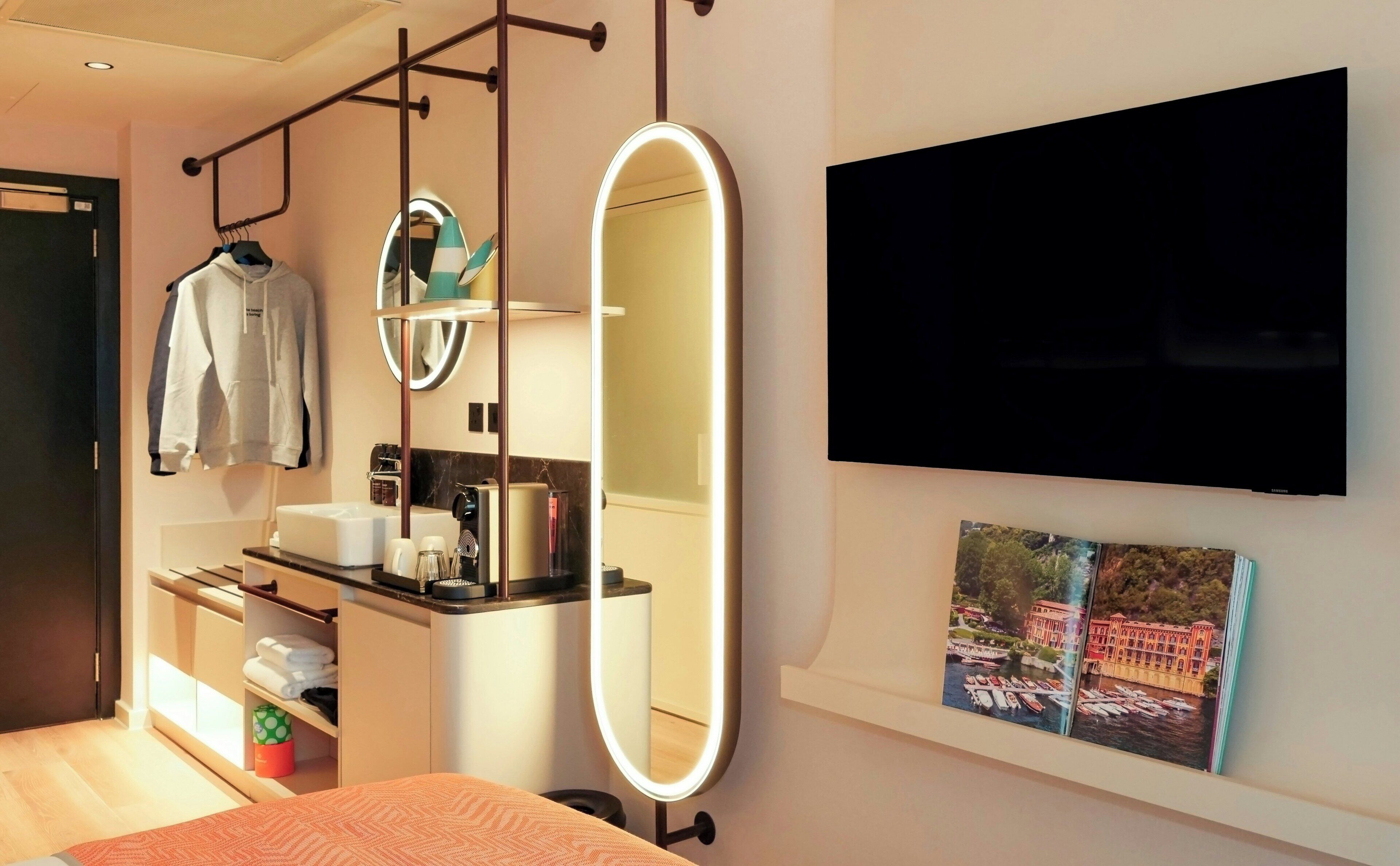 A modern hotel room with a bed, a vanity with a mirror, a flat-screen TV, and a picture of a scenic location.