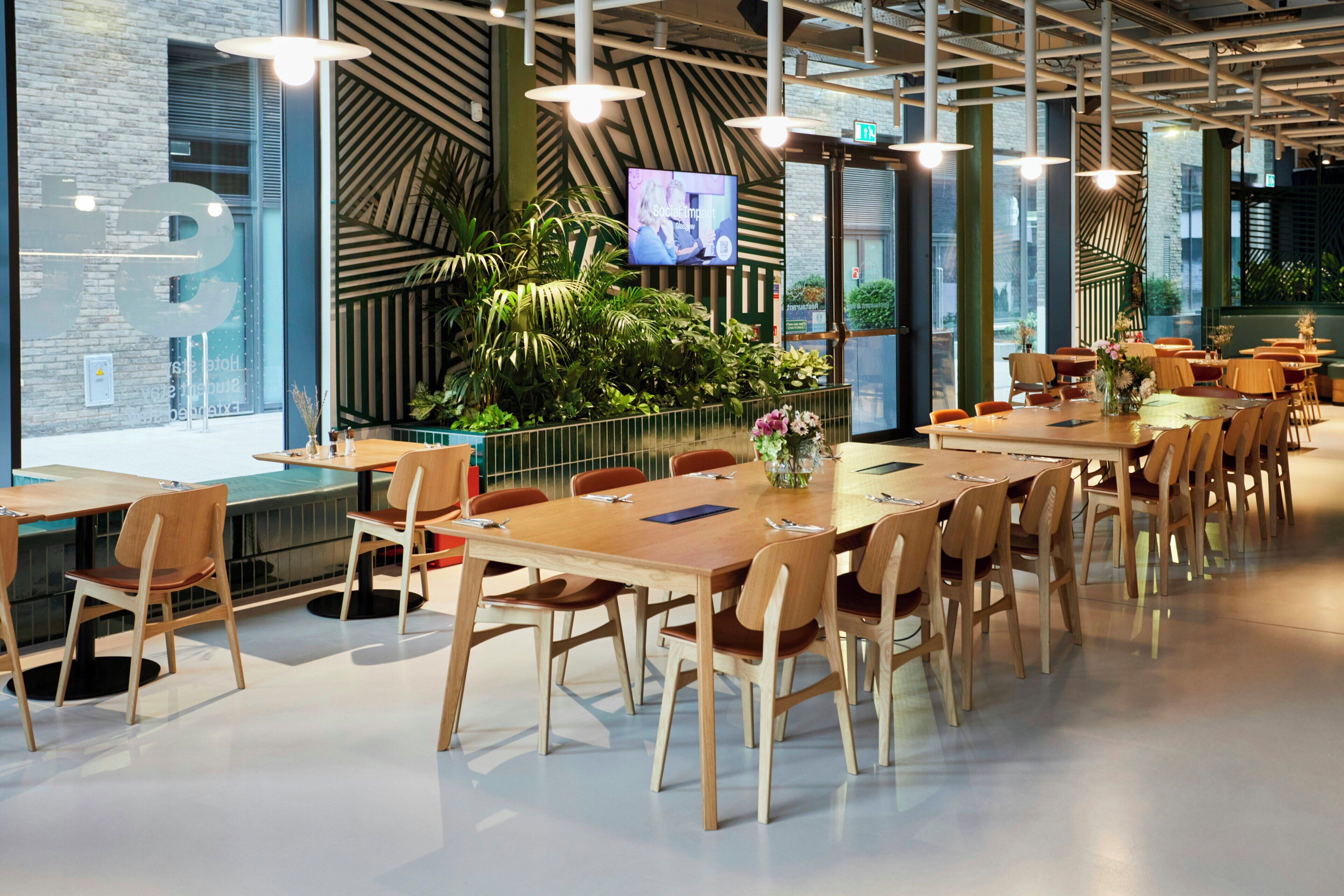 Un espace repas moderne avec de longues tables et des chaises en bois, un grand écran diffusant un programme et un mur vert orné d’un motif géométrique.