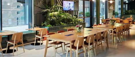 Un espace repas moderne avec de longues tables et des chaises en bois, un grand écran diffusant un programme et un mur vert orné d’un motif géométrique.