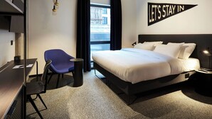 A hotel room with a bed, a desk, a chair, a small table, a wall-mounted TV, and a window with curtains.