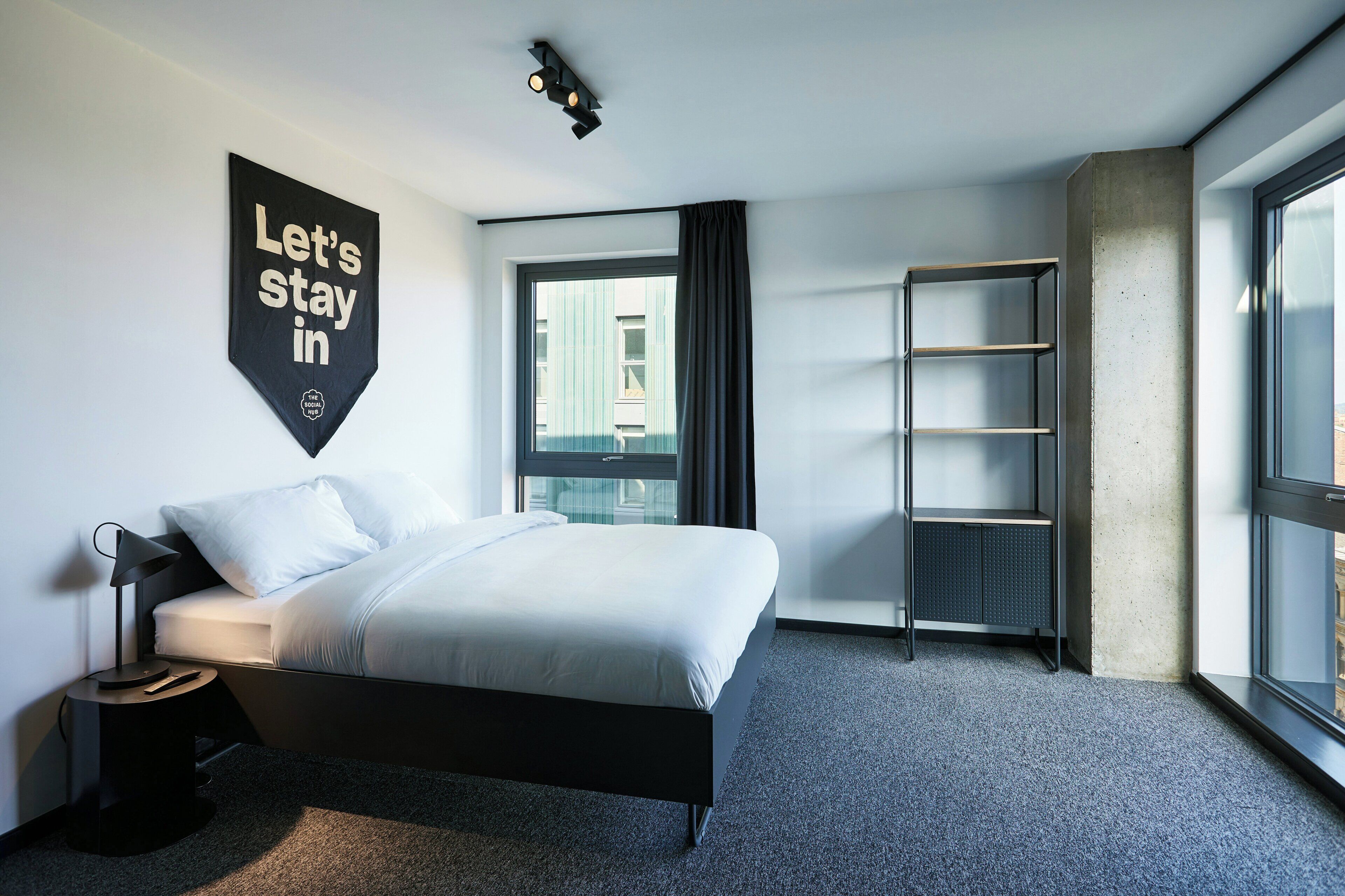 Een moderne hotelkamer met een bed, nachtkastje, een wandplank, een raam met gordijnen en een groot raam aan de rechterkant.