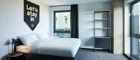Habitación de hotel moderna con cama, mesita de noche, estante en la pared, cortinas en la ventana y un ventanal grande a la derecha.