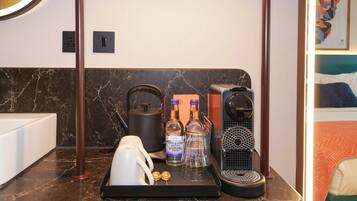 A hotel room with a marble countertop, a kettle, two bottles of whisky, a coffee maker, and a bed with a blue headboard.