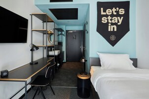 A modern hotel room with a desk, chair, bed, and a wall-mounted sign.