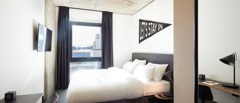 A modern hotel room with a large bed, a desk, a chair, and a view of the city.