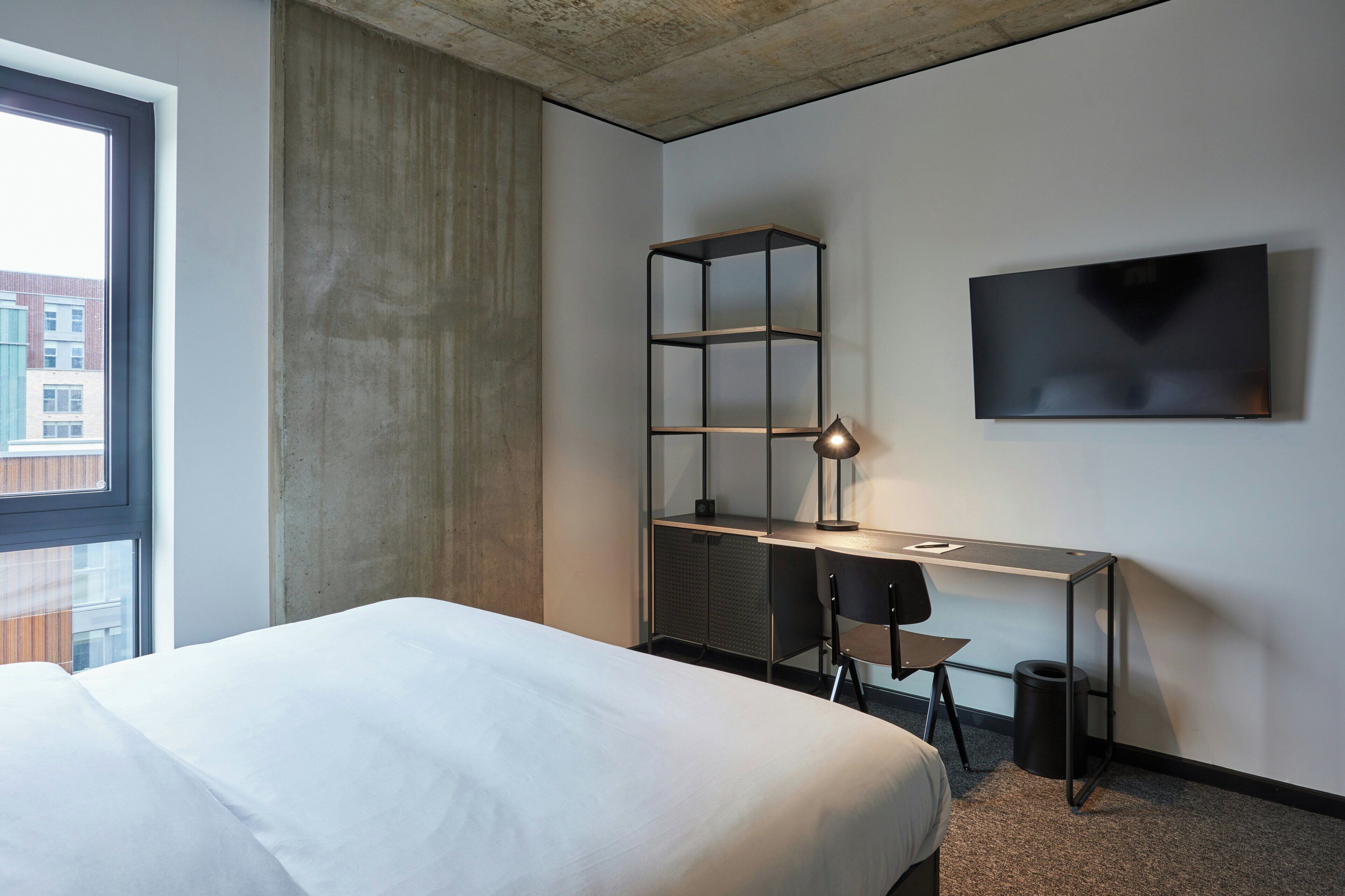Une chambre d’hôtel moderne, équipée d’un lit, d’un bureau avec une lampe, d’une télévision et offrant une vue sur les bâtiments environnants.