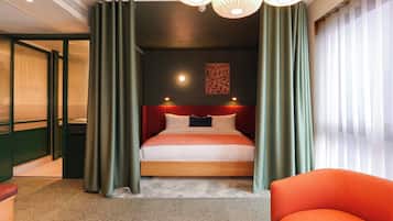 A hotel room with a bed, green curtains, a red headboard, a painting on the wall, and a bright ceiling light.