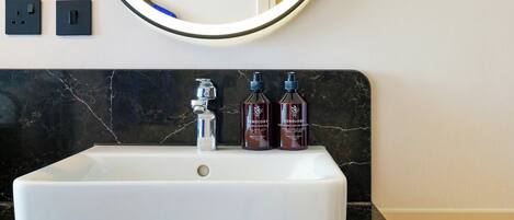 Un baño con un lavabo blanco, un espejo redondo y tres botellas de jabón de manos Zenology.