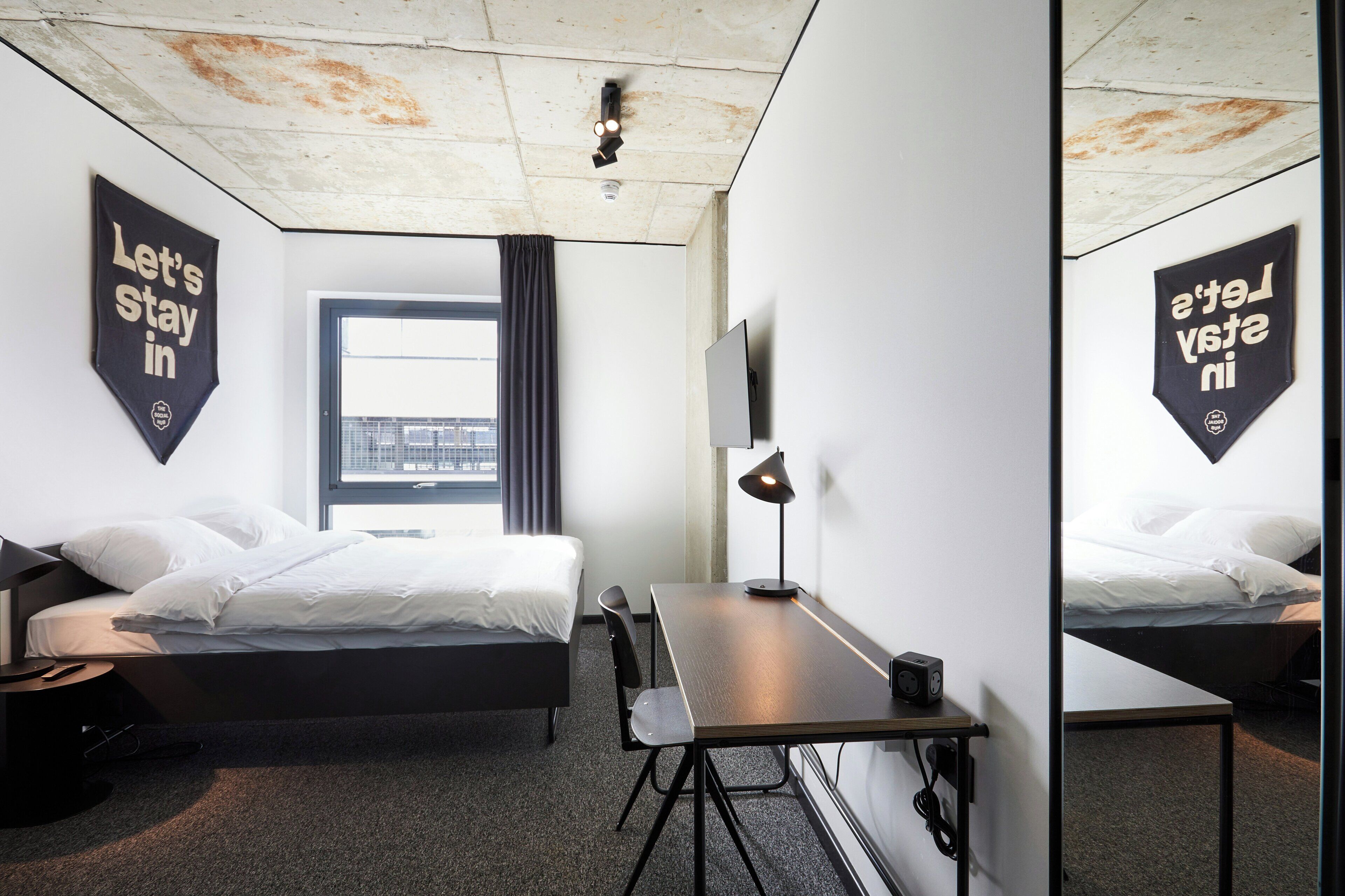 A hotel room with a bed, a desk, a chair, a lamp, and a window.