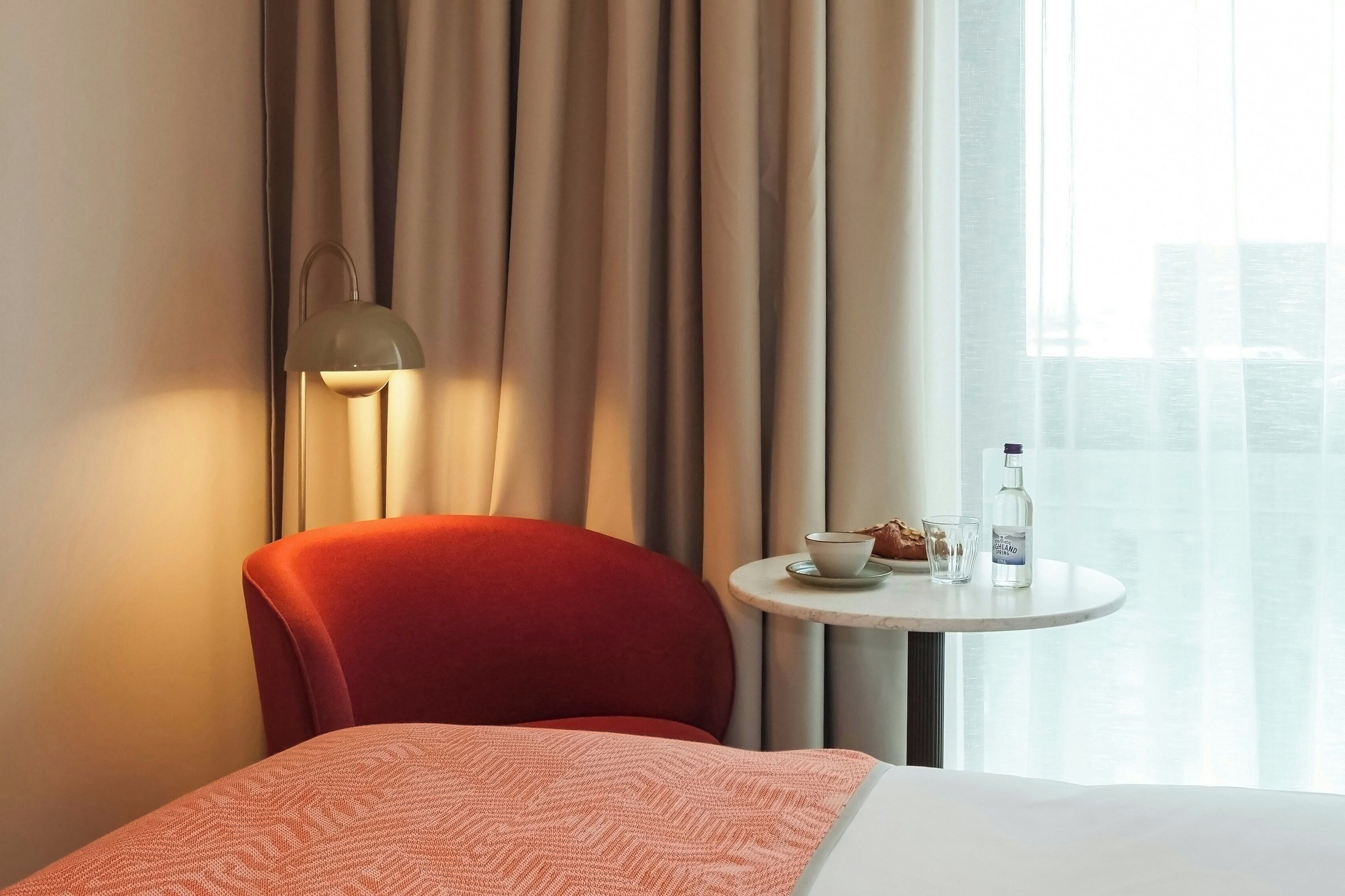A hotel room with a red chair, a bedside table with a cup and bottle, and a lamp.